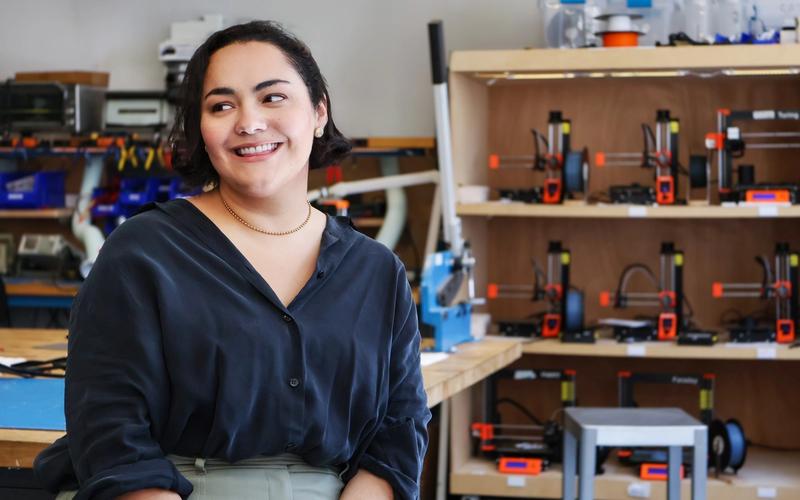 Young woman wearing a black shirt, seating and smiling in a makerspace. 3D printing tools in the background.
